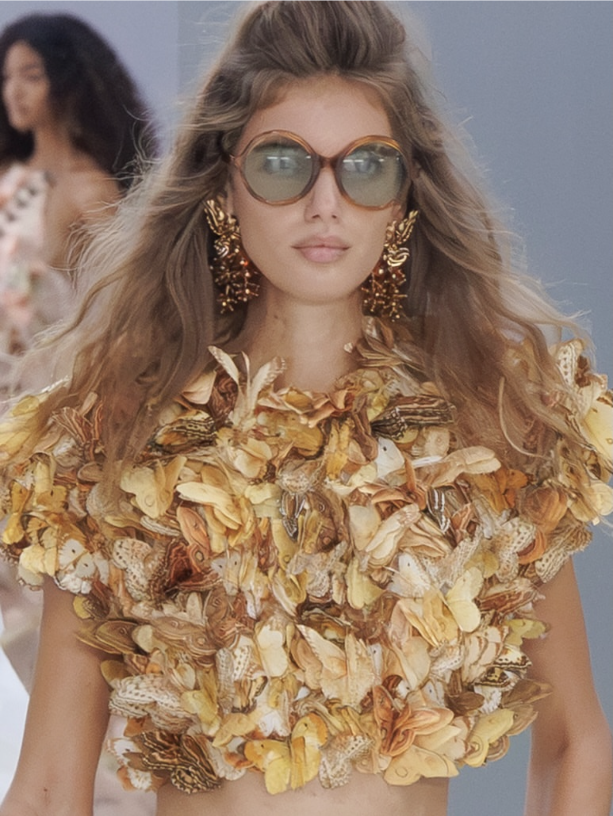 Modelo con lentes redondos grandes, aretes dorados y top de mariposas amarillas y naranjas 